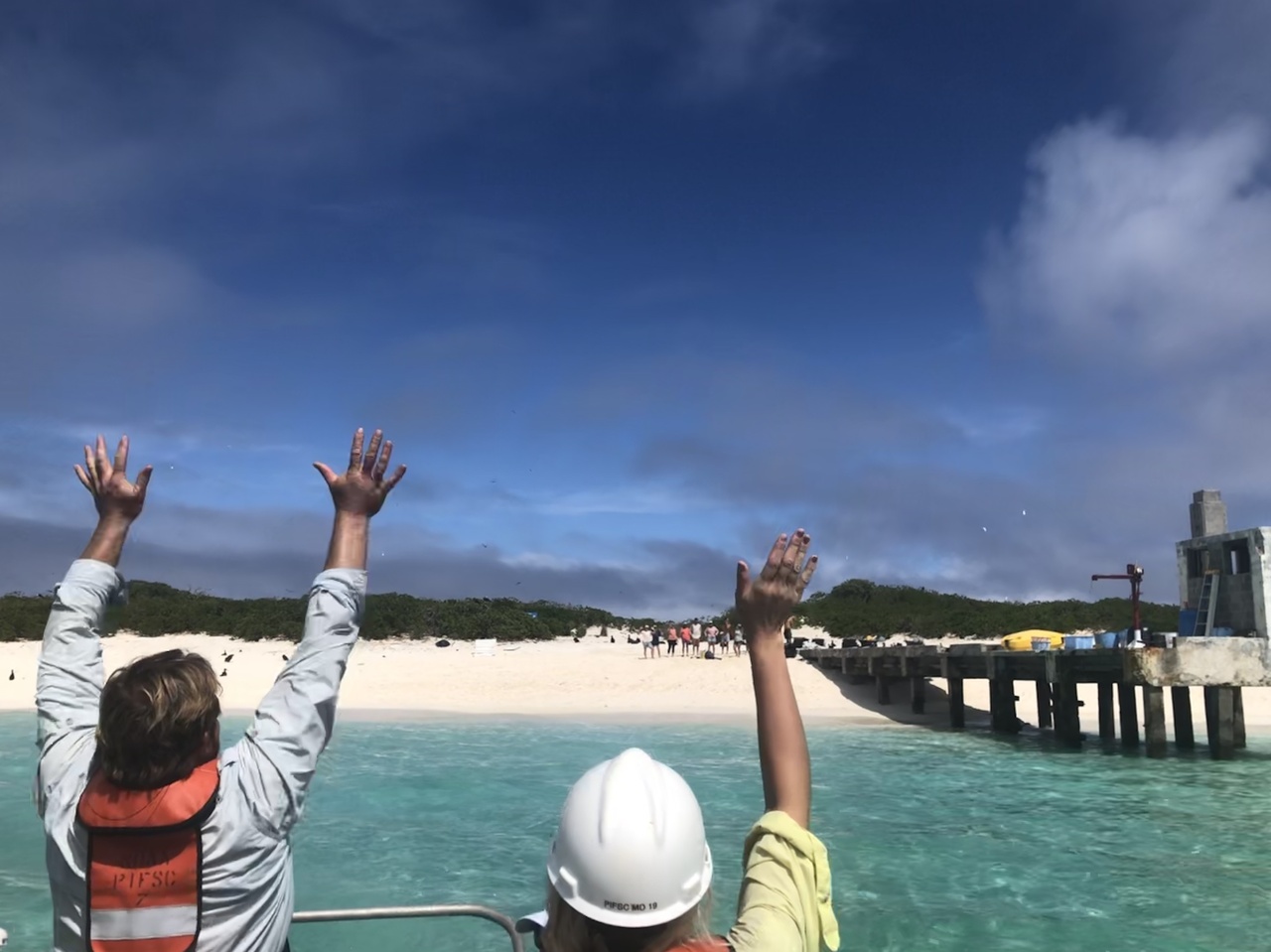 Two people wave from boat to field staff on beach at Kure.