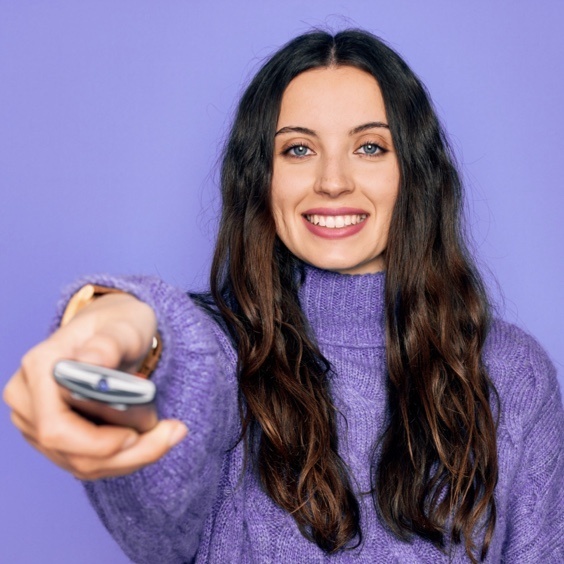 Woman wearing purple sweater smiling and holding and pointing television remot