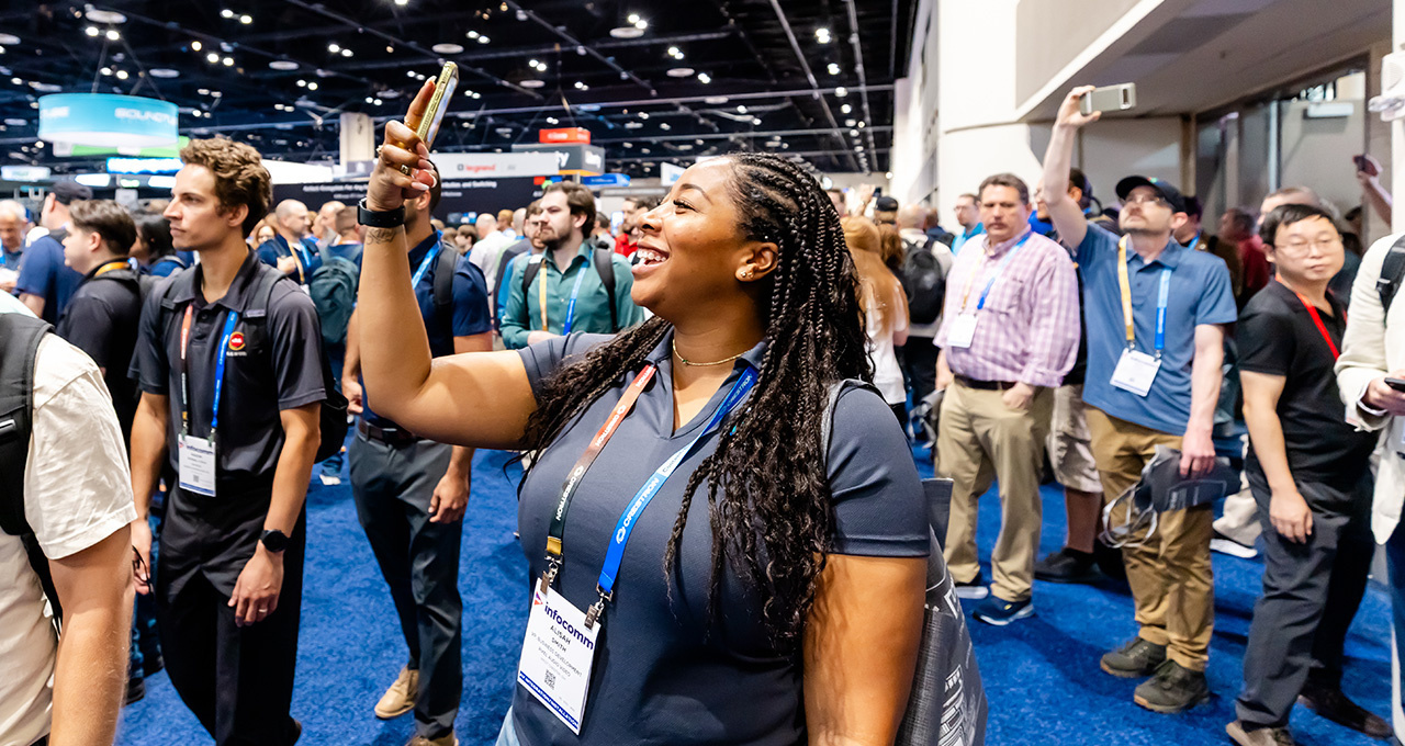 A woman wearing an event badge smiles while holding up her phone to take a photo or video in a crowded Expo 2025 trade show hall.
