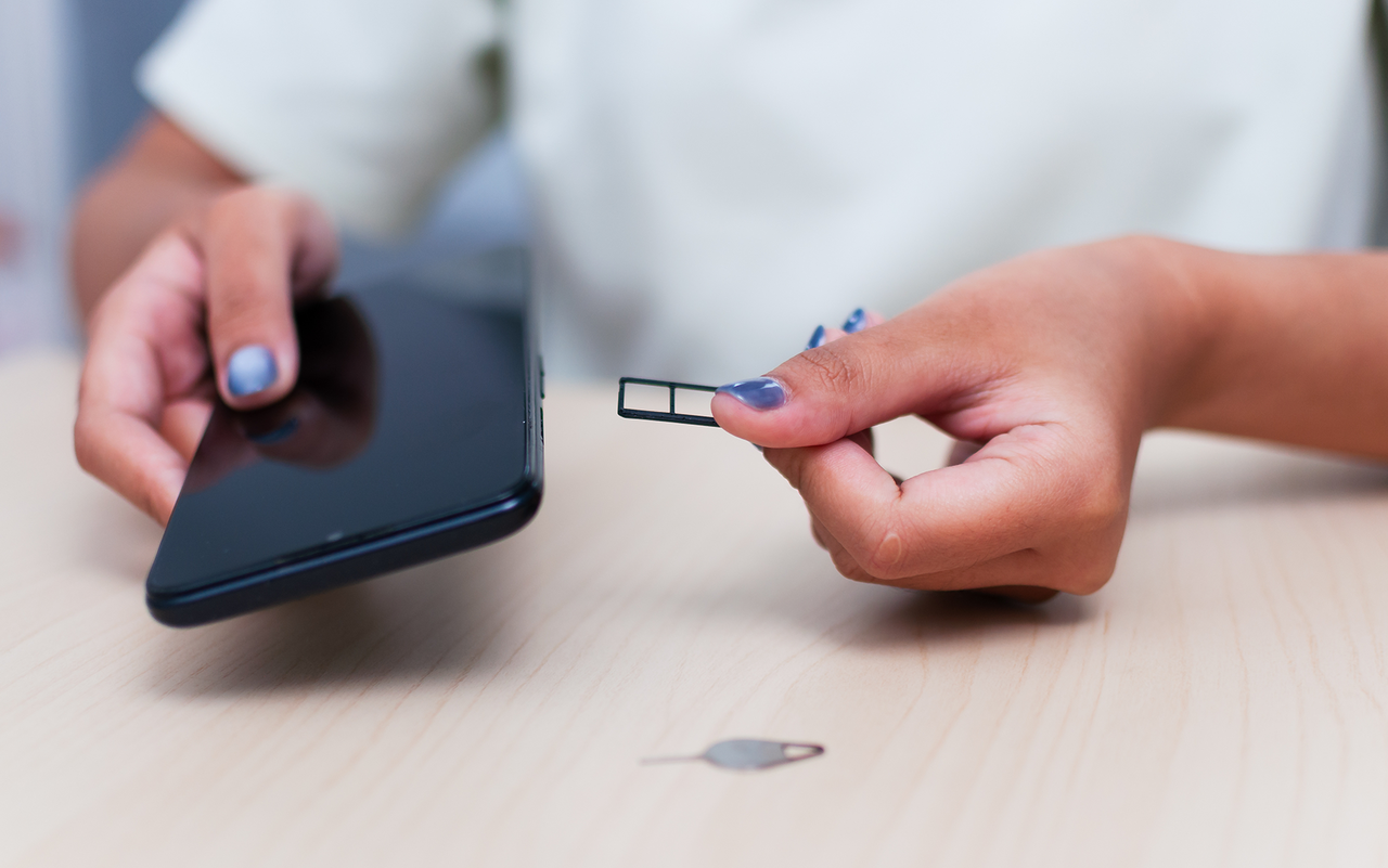 Person hält eine kleine Nano-SIM-Karte zwischen den Fingern und ein dunkles Smartphone in der anderen Hand.